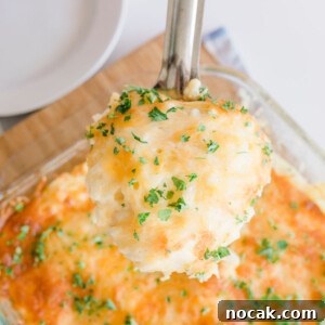 Cracker Barrel Hashbrown Casserole Copycat Recipe 13 A serving spoon holds a portion of cheesy mashed potatoes reminiscent of a cracker barrel hashbrown casserole, topped with melted cheese and chopped parsley. The dish rests in a glass baking dish on a wooden board, with an empty white plate nearby.