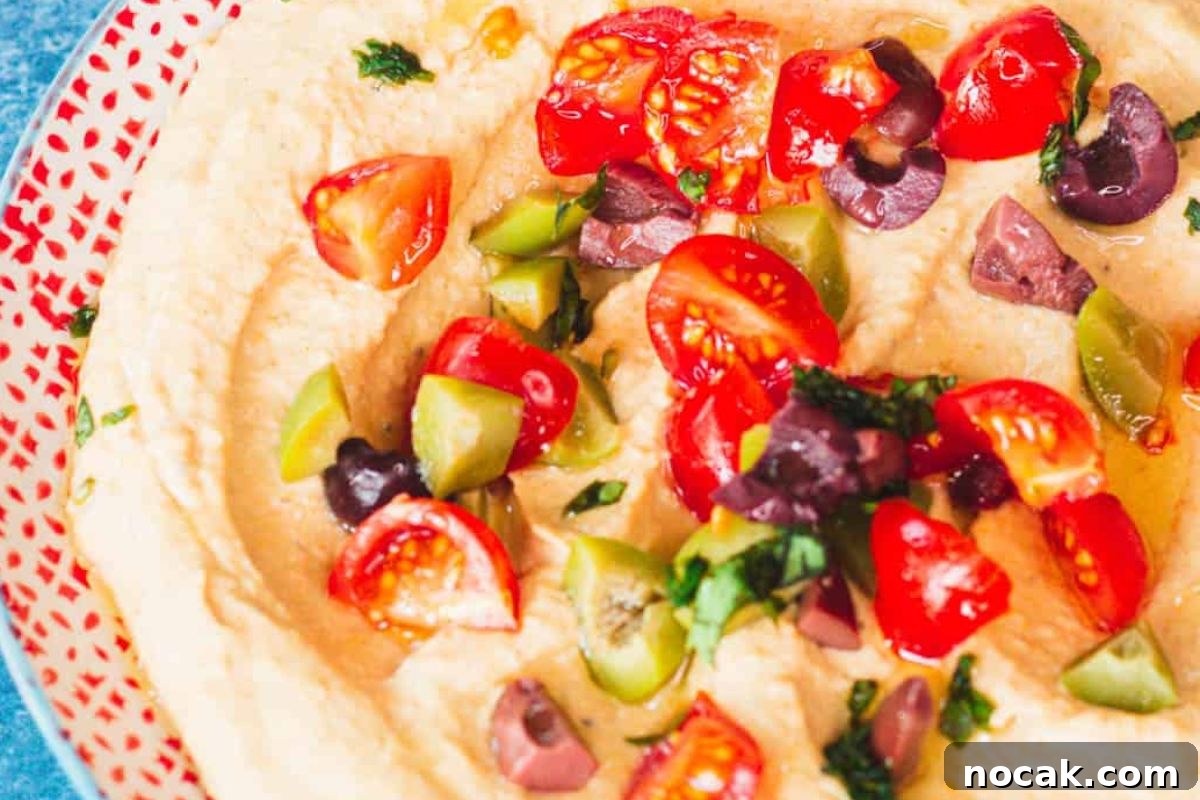 35 Easter Appetizers to Kick Off Your Holiday Feast 26 Hummus topped with tomatoes, olives, and herbs served on a patterned plate as an Easter appetizer.