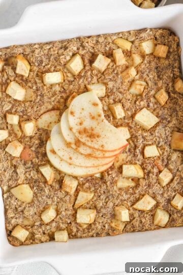 Baked apple oatmeal topped with apple slices in a square white baking dish with apples and syrup on the side.