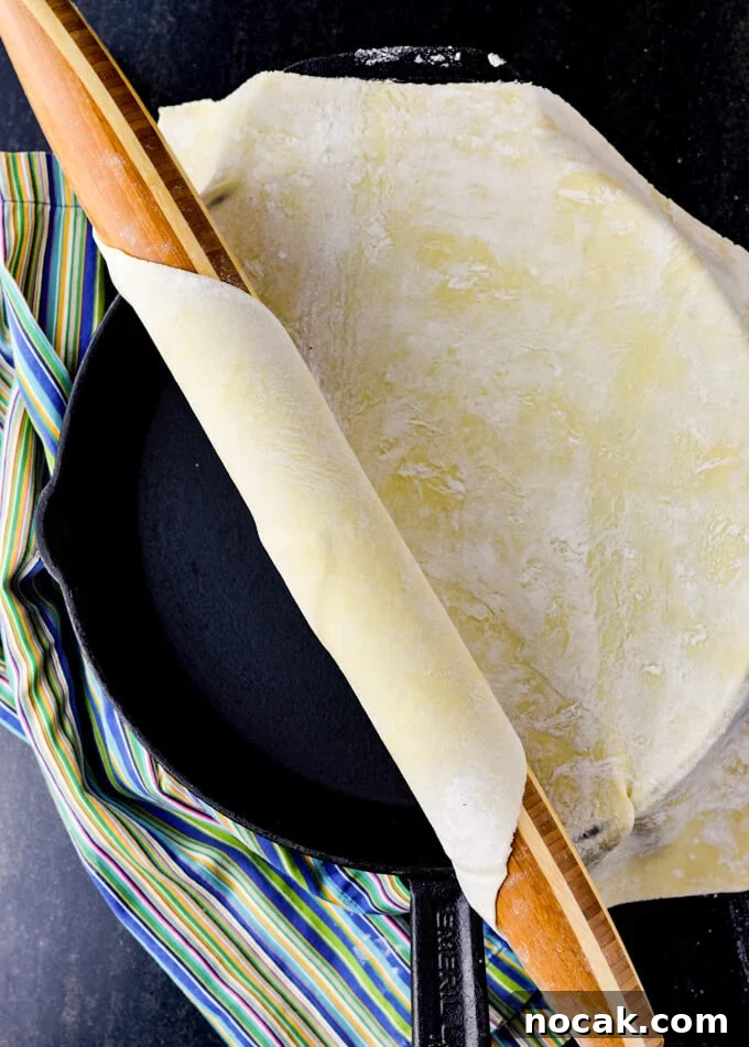 Rustic Apple Galette with Flaky Puff Pastry 3 A rolling pin is rolling out a sheet of puff pastry onto a large black cast iron skillet. A blue striped napkin sits beside the skillet.