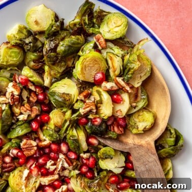 Roasted Brussels Sprouts and Pomegranate Salad with Citrus Vinaigrette 13 The brussels sprouts have been placed into a white, oval-shaped serving dish with a blue border. Pomegranate seeds and chopped pecans have been added on top. A wooden serving spoon is shown scooping the brussels sprouts.