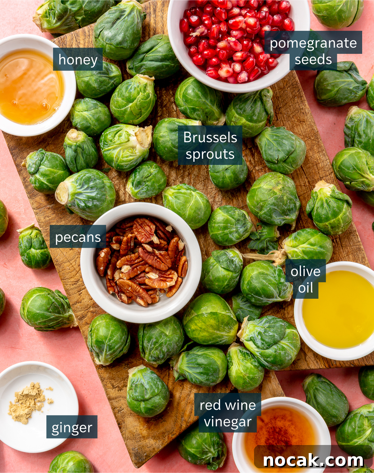 Roasted Brussels Sprouts and Pomegranate Salad with Citrus Vinaigrette 3 Ingredients for roasted pomegranate brussels sprout slaw sit in a variety of bowls and on a wooden chopping block. All sit on a light pink background.