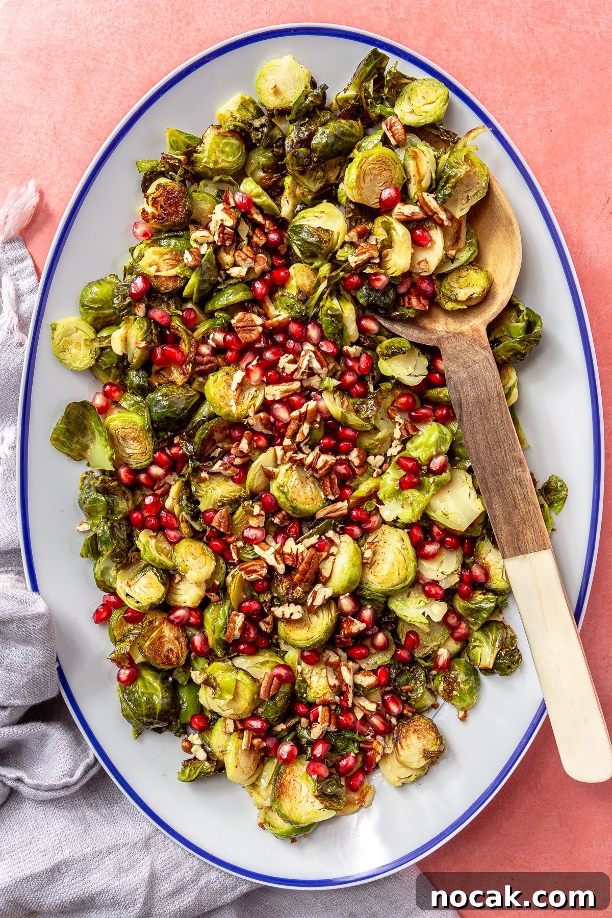Roasted Brussels Sprouts and Pomegranate Salad with Citrus Vinaigrette 8 The brussels sprouts have been placed into a white, oval-shaped serving dish with a blue border. Pomegranate seeds and chopped pecans have been added on top. A wooden serving spoon is shown scooping the brussels sprouts.