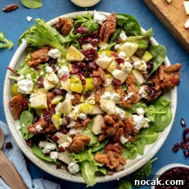 Mixed green salad garnished with goat cheese, candied pecans, pears, and dried cranberries in a large, white bowl on a blue table surrounded by small bowls of garnish, diced pears, salad dressing, and salad servers.