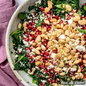 The remaining ingredients, including pomegranate seeds and chopped almond, have been added to the top of the kale. A purple dishcloth sits to the side.