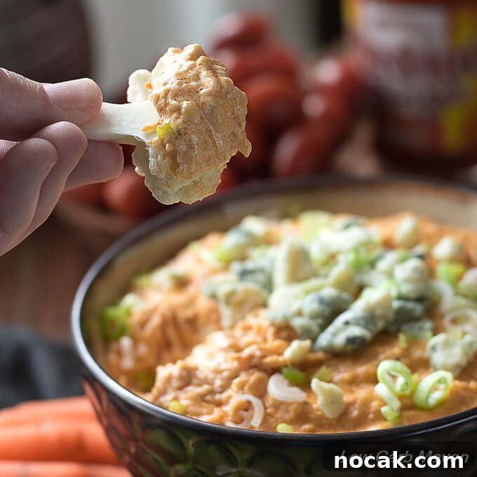 Keto Buffalo Chicken Dip Recipe — Low Carb, Creamy & Spicy 5 Low Carb Buffalo Chicken Dip topped with blue cheese and scallions in a brown bowl surrounded by vegetables. A piece of cauliflower is shown after being dipped into the hot appetizer.
