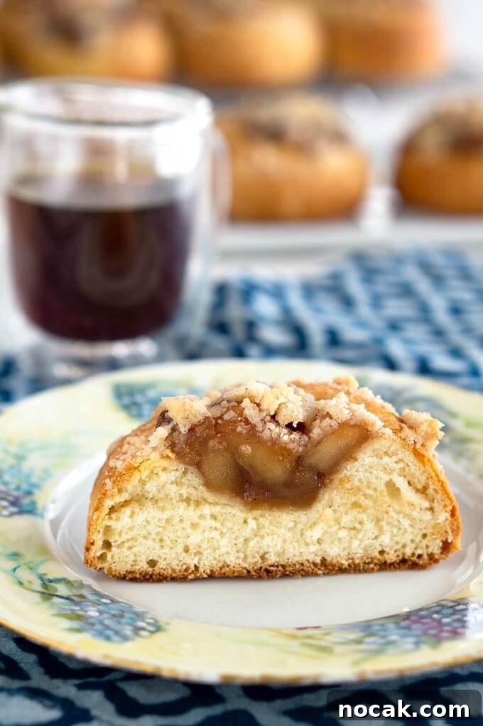 Homemade Apple Kolache Recipe with Video Tutorial 14 Pastry roll filled with apple filling cut in half sitting on a floral plate. A cup of coffee and more rolls sit behind the plate.