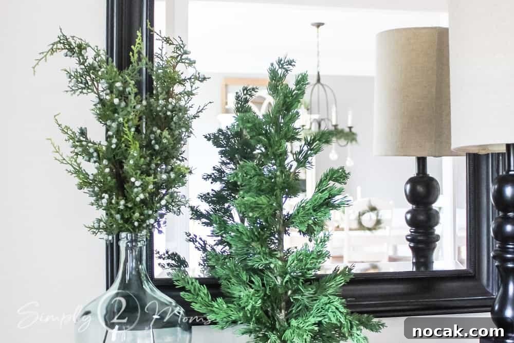 faux juniper stems with berries in glass bottle neck jar next to mini faux tree on table in front of mirror