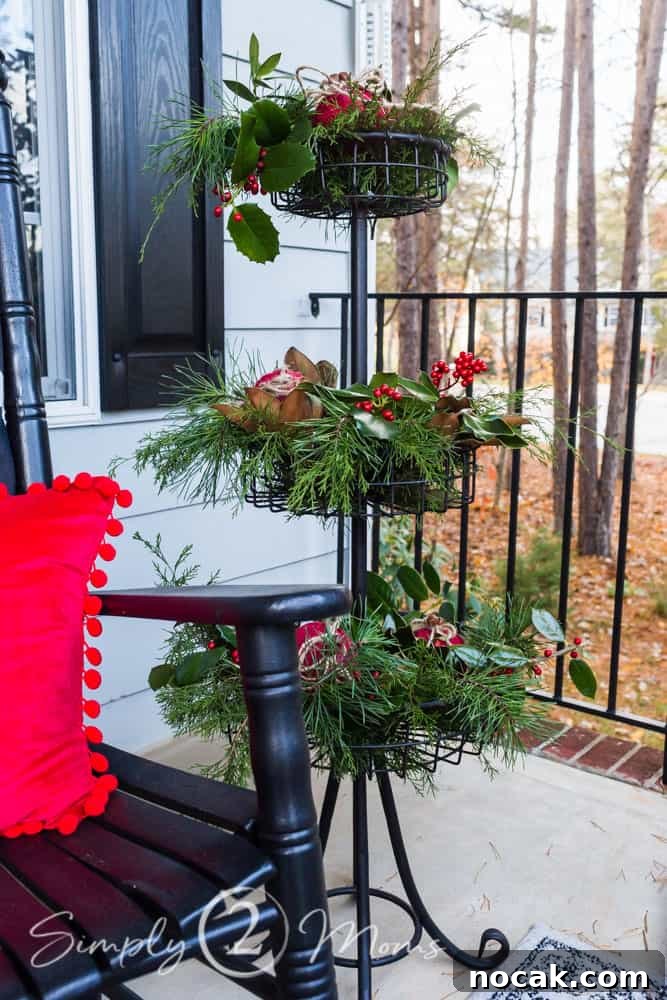 iron tiered plant stand on a front porch filled with assorted fresh greenery, red berries, and large fabric covered ornaments