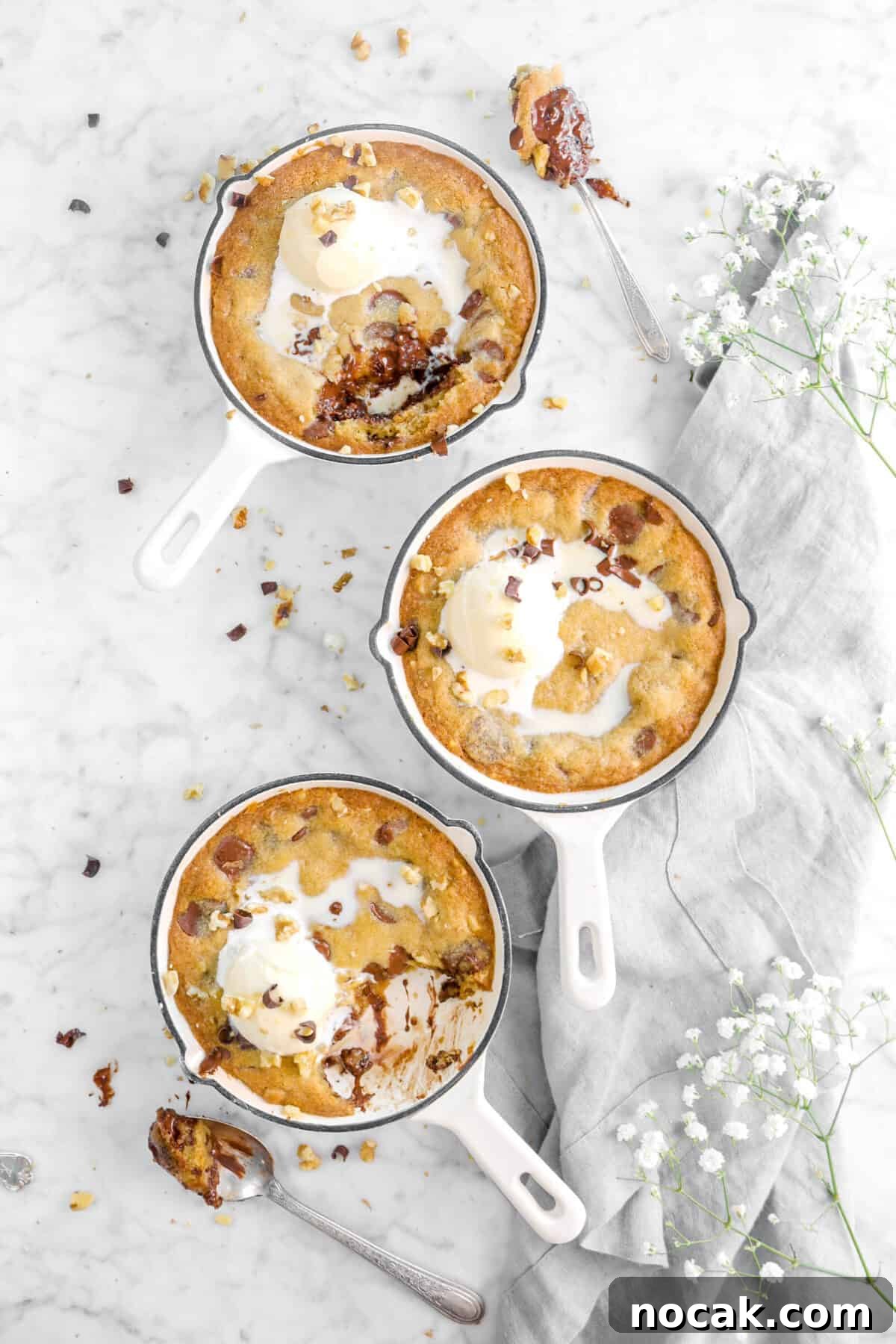 overhead shot of three skillet cookies with a grey napkin, white flowers, two spoonfuls of cookie