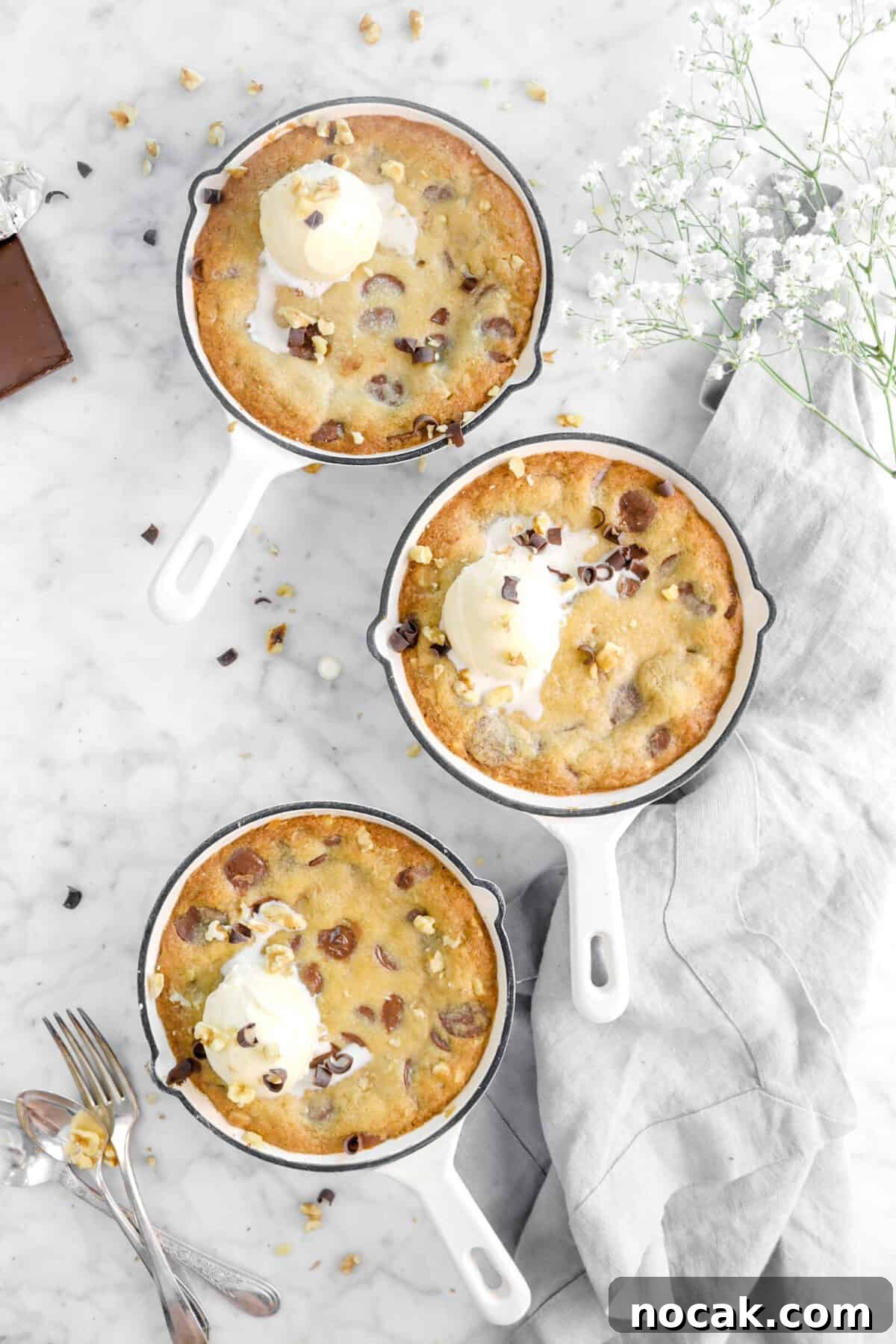 three white skillets of cookie with ice cream, chocolate curls, white flowers, and utensils