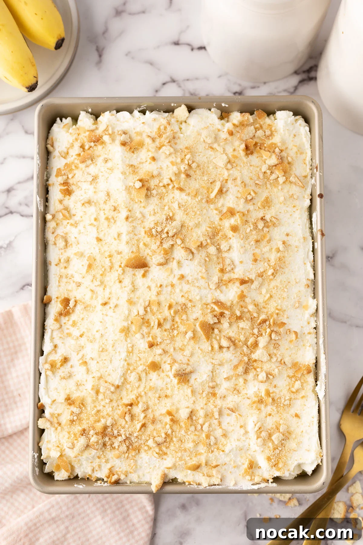 Creamy Banana Pudding Poke Cake Recipe for Dessert Fans 10 Homemade whipped cream frosting spread over top of cake with crumbled vanilla wafers in the baking pan.