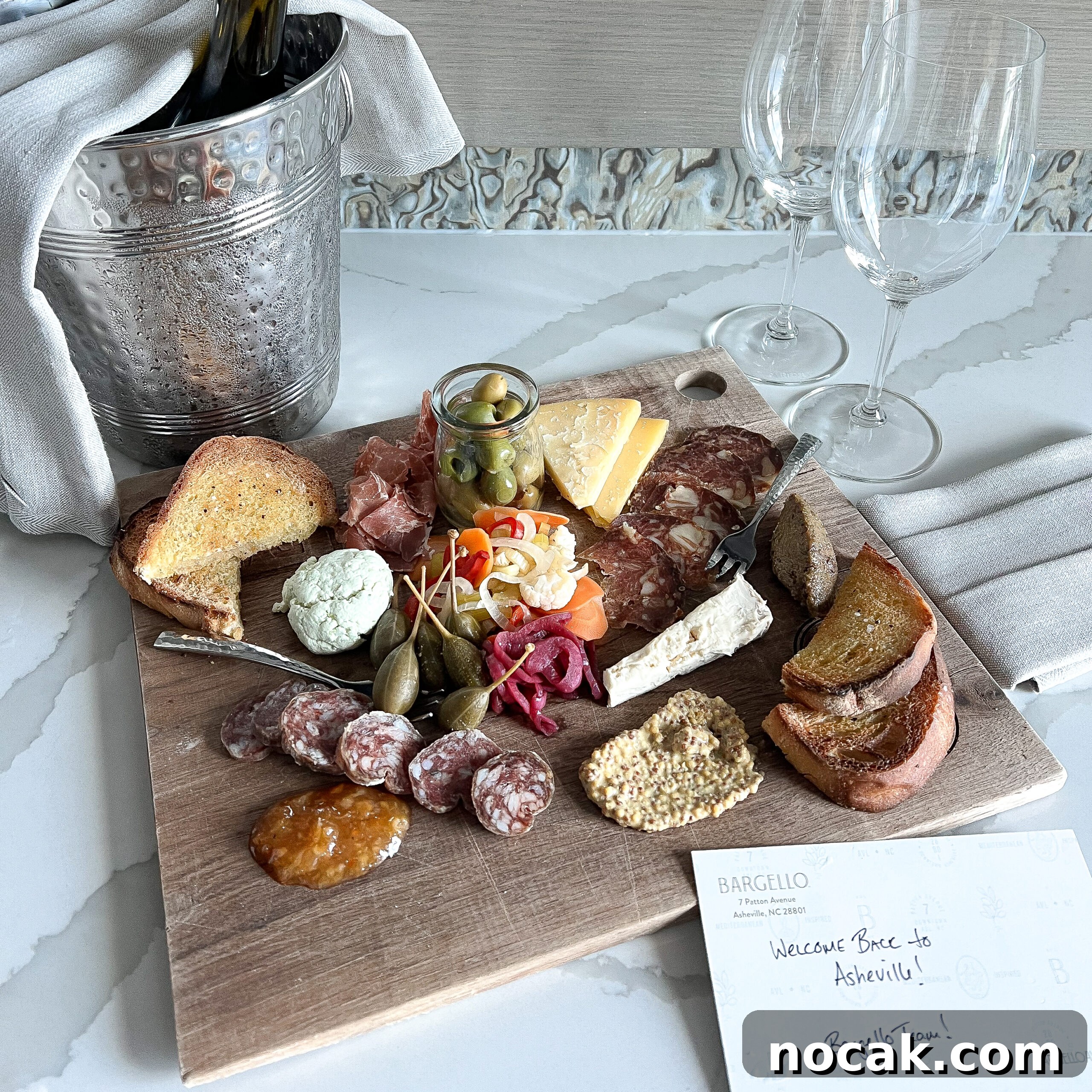 A wooden charcuterie board with cheeses, olives, and toasted bread beside two wine glasses.
