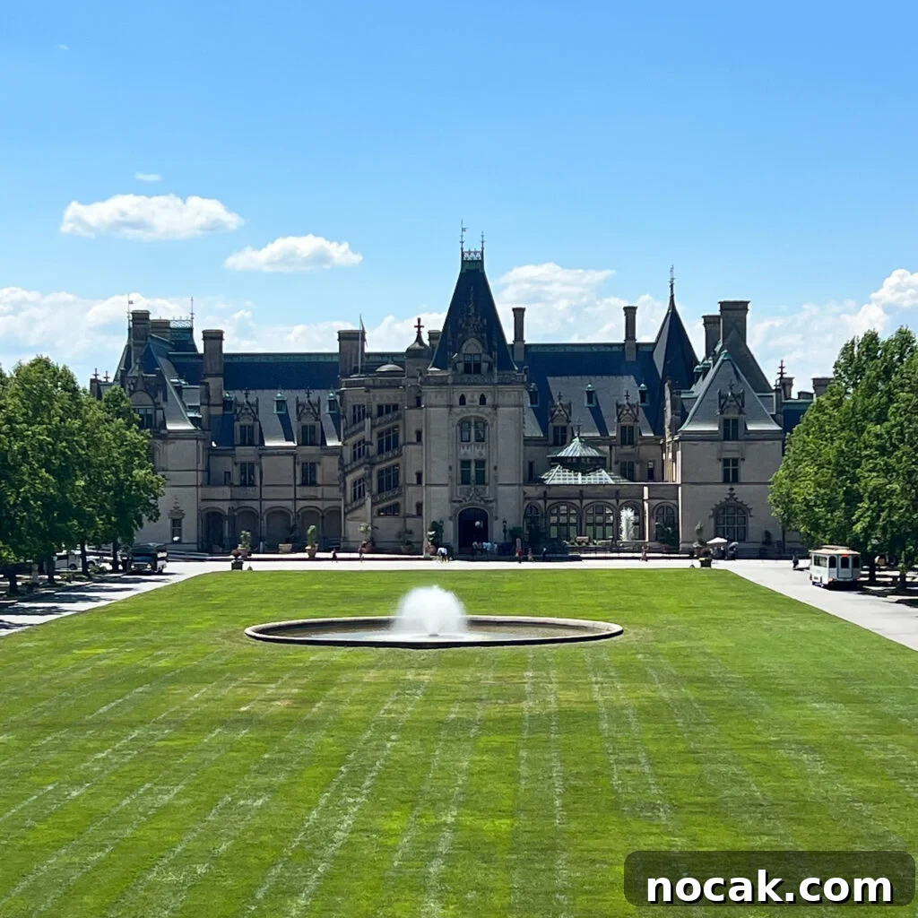 A large romantic historic mansion with a central fountain on a green lawn, surrounded by trees under a bright blue sky with scattered clouds.