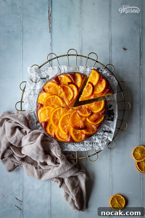 Sticky Orange Glaze Cake Recipe with Zesty Citrus Flavor 2 A distanced flat lay of a sticky orange cake with a slice cut out of it on a round cooling rack over a cool grey wood effect backdrop. There are also dried orange slices and a light brown linen napkin in the background.