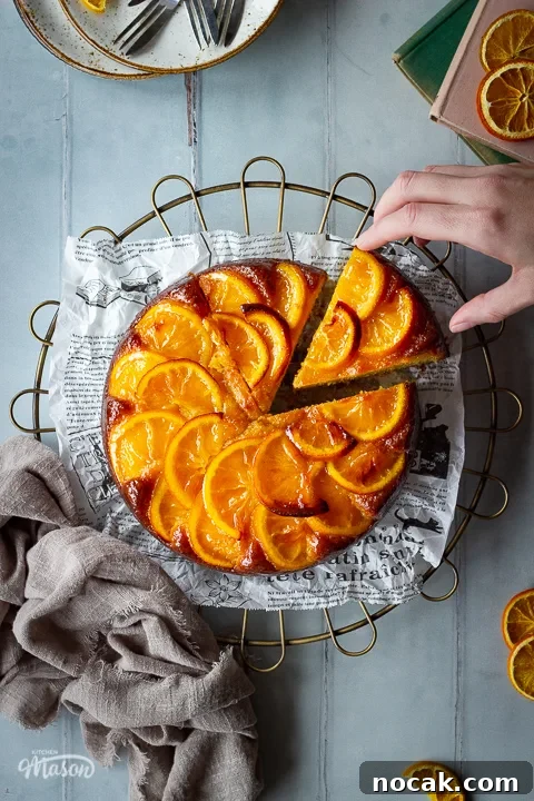 Sticky Orange Glaze Cake Recipe with Zesty Citrus Flavor 6 A sticky orange cake with a slice cut out of it and a hand reaching in on a round cooling rack over a cool grey wood effect backdrop. There are also dried orange slices, white rustic plates with dessert forks, two stacked books and a light brown linen napkin in the background.
