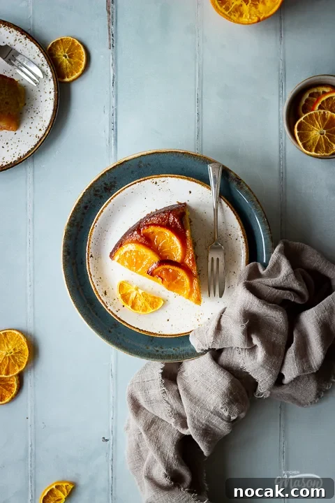 Sticky Orange Glaze Cake Recipe with Zesty Citrus Flavor 7 Flay lay view of a slice of sticky orange cake on two stacked plates with a fork. Set over a cool grey wood effect backdrop, there are also dried orange slices, another plate with cake, a small pot filled with orange slices and a light brown linen napkin in the background.