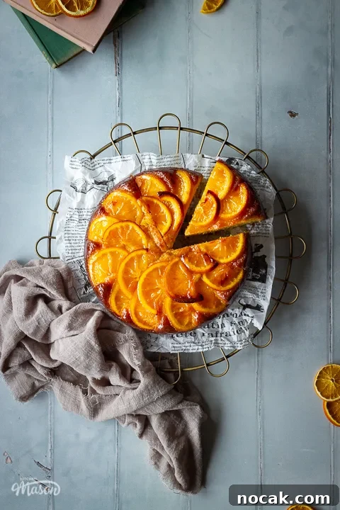 Sticky Orange Glaze Cake Recipe with Zesty Citrus Flavor 8 A distanced flat lay of a sticky orange cake with a slice cut out of it on a round cooling rack over a cool grey wood effect backdrop. There are also dried orange slices, two stacked books and a light brown linen napkin in the background.