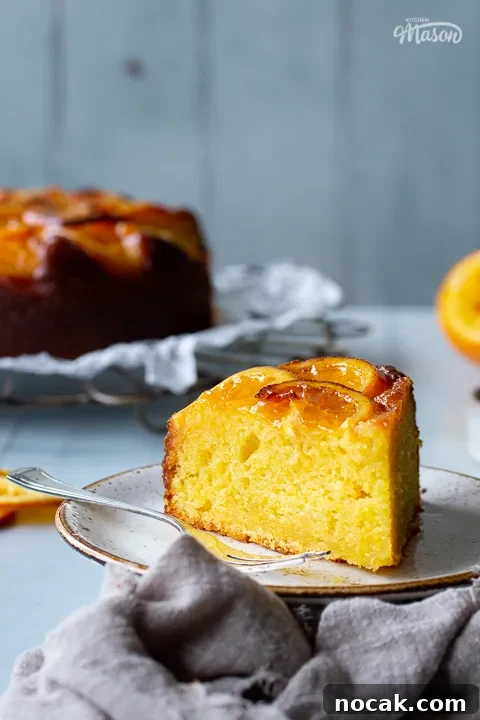 Sticky Orange Glaze Cake Recipe with Zesty Citrus Flavor 9 A front view of a slice of sticky orange cake on a plate with a fork positioned to the right of the shot. In the background there is the remaining sticky orange cake on a round cooling rack, half a squeezed orange, some dried orange slices and a light brown linen napkin. All set over a cool grey wood effect backdrop.