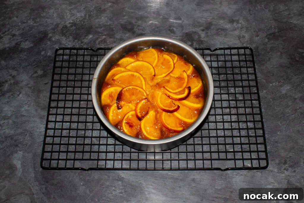 Sticky Orange Glaze Cake Recipe with Zesty Citrus Flavor 17 Baked sticky orange cake topped with orange syrup in a round baking tin on a cooling rack on a kitchen worktop.