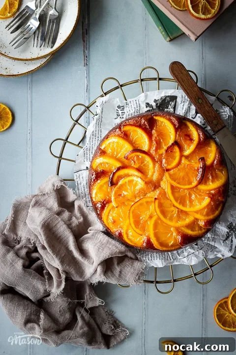 Sticky Orange Glaze Cake Recipe with Zesty Citrus Flavor 18 A whole sticky orange cake on a round cooling rack positioned to the right side over a cool grey wood effect backdrop. There are also dried orange slices, white rustic plates with dessert forks, two stacked books and a light brown linen napkin in the background.