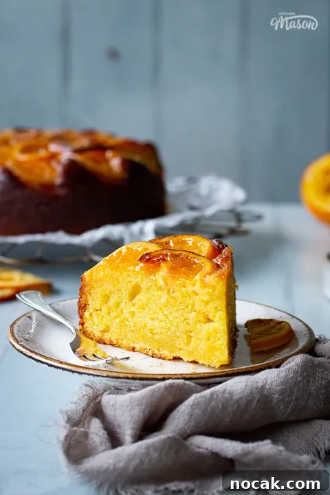 Sticky Orange Glaze Cake Recipe with Zesty Citrus Flavor 19 A front view of a slice of sticky orange cake on a plate with a fork positioned to the centre of the shot. In the background there is the remaining sticky orange cake on a round cooling rack, half a squeezed orange, some dried orange slices and a light brown linen napkin. All set over a cool grey wood effect backdrop.