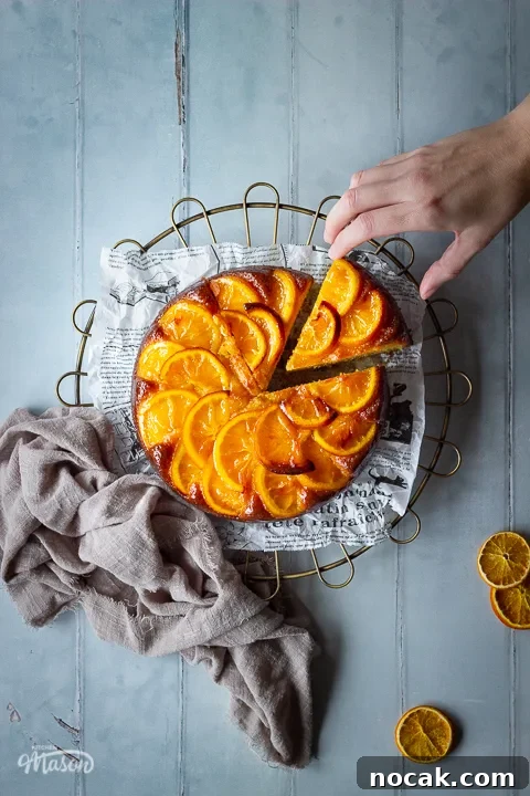 Sticky Orange Glaze Cake Recipe with Zesty Citrus Flavor 20 A distanced flat lay of a sticky orange cake with a slice cut out of it on a round cooling rack with a hand reaching in over a cool grey wood effect backdrop. There are also dried orange slices and a light brown linen napkin in the background.
