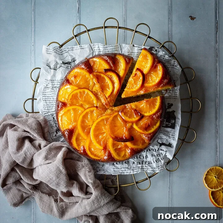 A flat lay of a sticky orange cake with a slice cut out of it on a round cooling rack over a cool grey wood effect backdrop. There are also dried orange slices and a light brown linen napkin in the background.