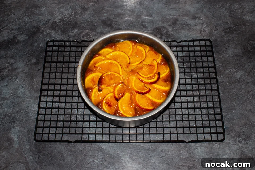 Sticky Orange Glaze Cake Recipe with Zesty Citrus Flavor 31 Baked sticky orange cake topped with orange syrup in a round baking tin on a cooling rack on a kitchen worktop.
