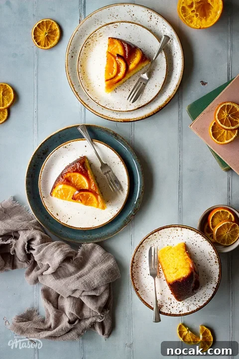 Sticky Orange Glaze Cake Recipe with Zesty Citrus Flavor 5 A flat lay view of 3 plates, each with a slice of sticky orange cake and a fork on them, set over a cool grey wood effect backdrop. There are also dried orange slices, two stacked books, half a squeezed orange and a light brown linen napkin in the background.