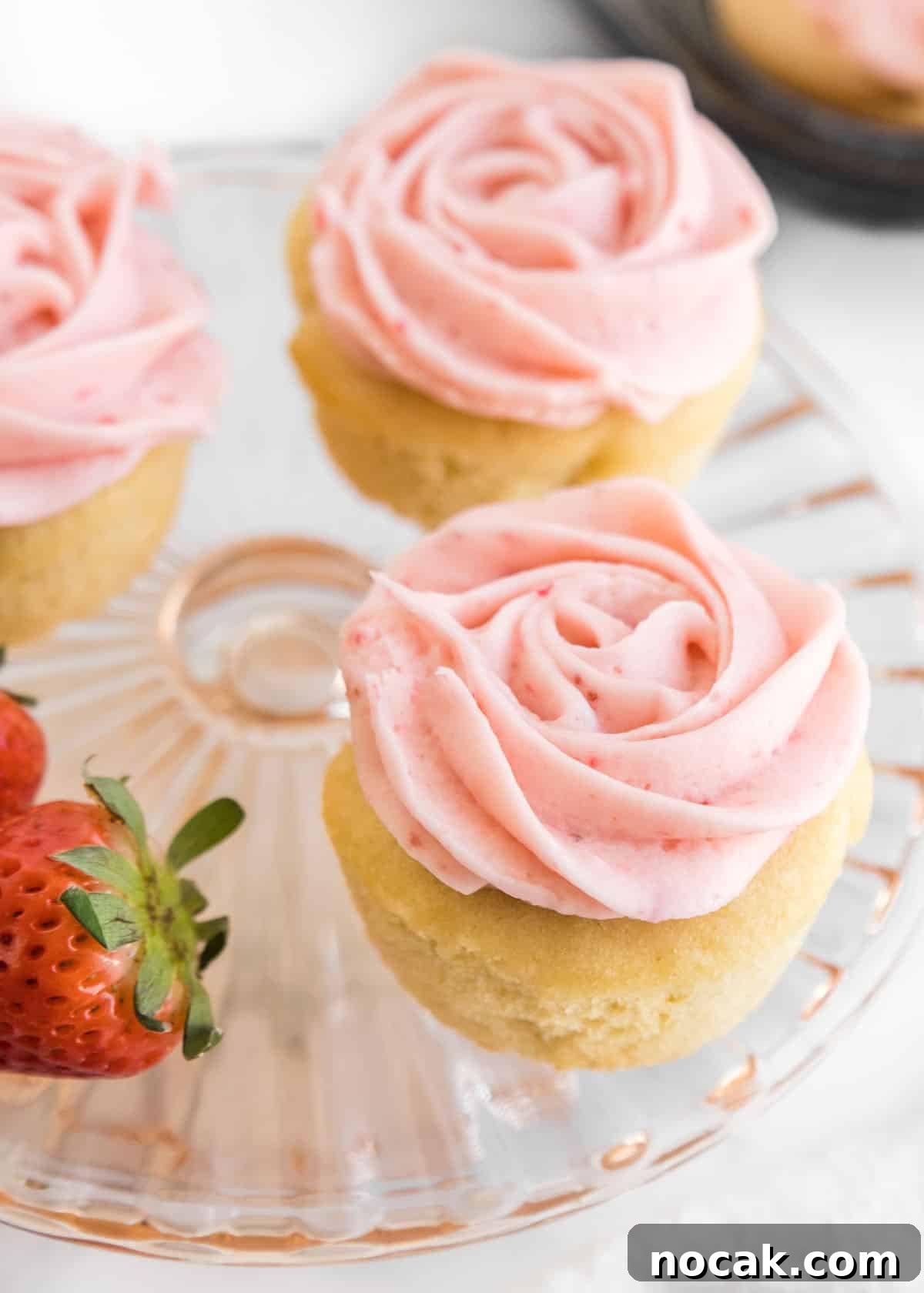 Strawberry Buttercream Frosting Recipe — Smooth, Fresh Flavor 2 A close-up photo of three cupcakes and two whole strawberries on a small, glass cake stand.