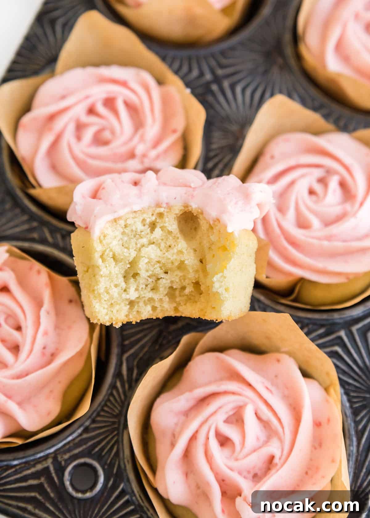 Strawberry Buttercream Frosting Recipe — Smooth, Fresh Flavor 11 A close up photo of one cupcake with a bite taken out of it placed on top of a muffin tin filled with the strawberry buttercream cupcakes.