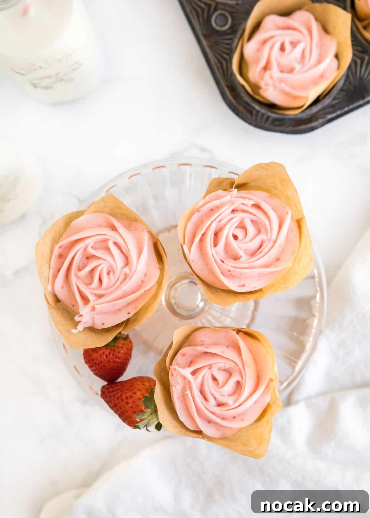 Strawberry Buttercream Frosting Recipe — Smooth, Fresh Flavor 12 Overhead shot of three strawberry buttercream cupcakes and two whole strawberries on a small, glass cake stand. The top left and right corners are accented by a glass of milk and a cupcake in it's parchment liner in the muffin tin.