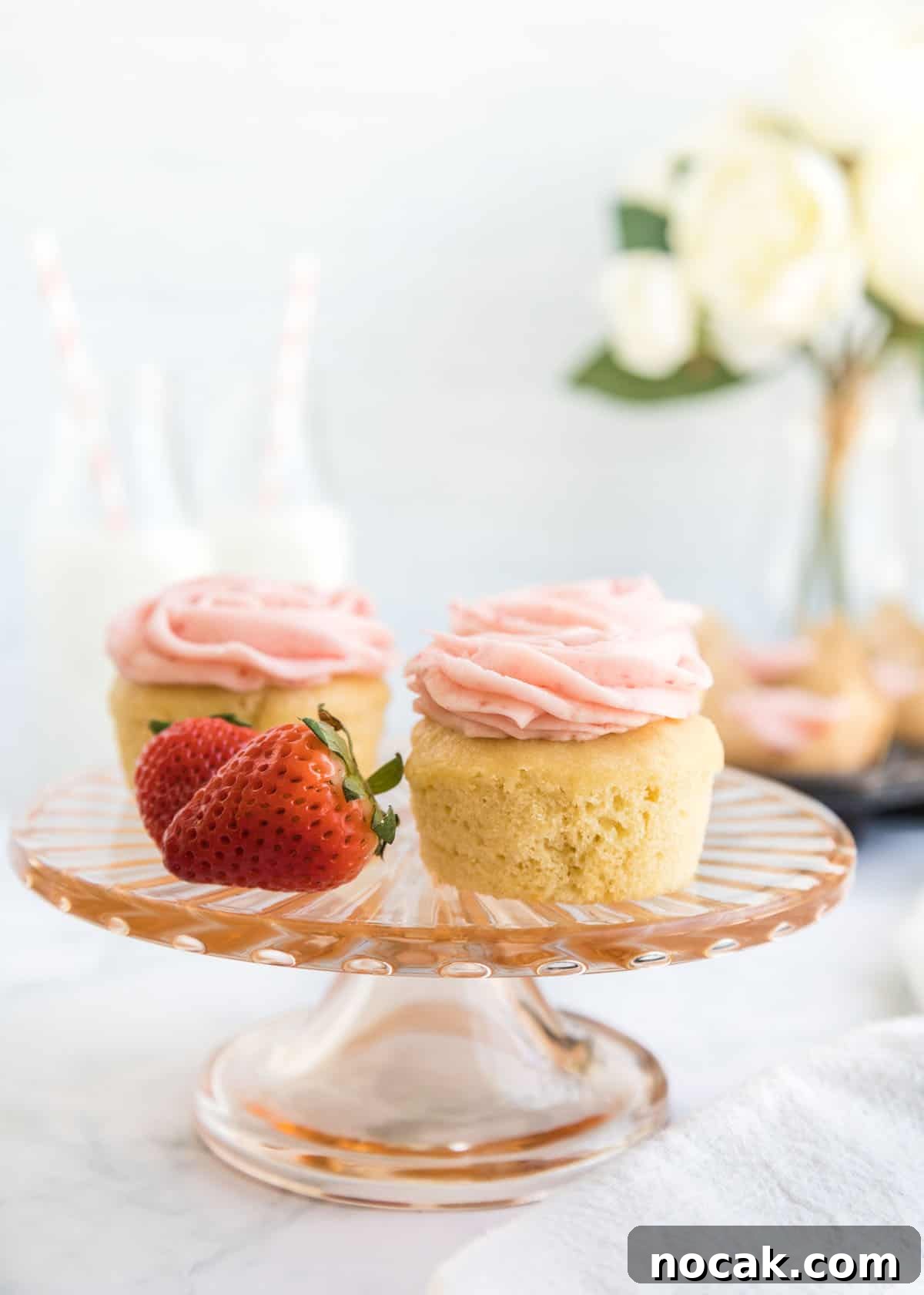 Strawberry Buttercream Frosting Recipe — Smooth, Fresh Flavor 13 Close-up photo of two cupcakes and two whole strawberries on a small, glass cake stand with two milk jars and a small bouquet blurred in the background.