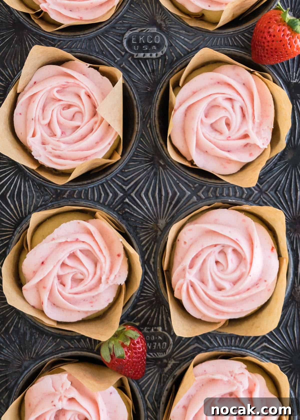 Strawberry Buttercream Frosting Recipe — Smooth, Fresh Flavor 3 Overhead shot of fully baked strawberry butter cream cupcakes in a cupcake tin with full strawberries accenting the edges of the tin.