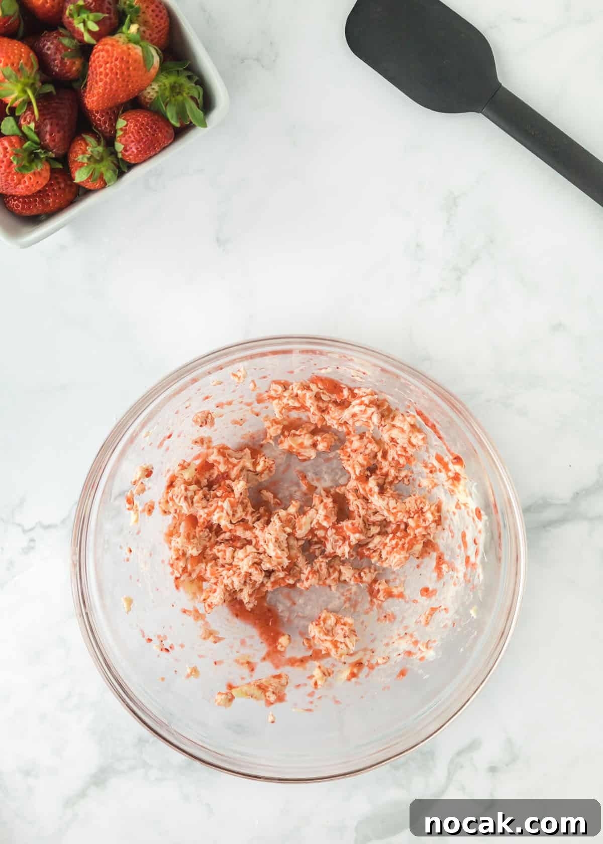 Strawberry Buttercream Frosting Recipe — Smooth, Fresh Flavor 6 Overhead photo of pureed strawberries with one full stick of butter and vanilla mixed together. The upper left and right corners are accented with whole strawberries and a black spatula.