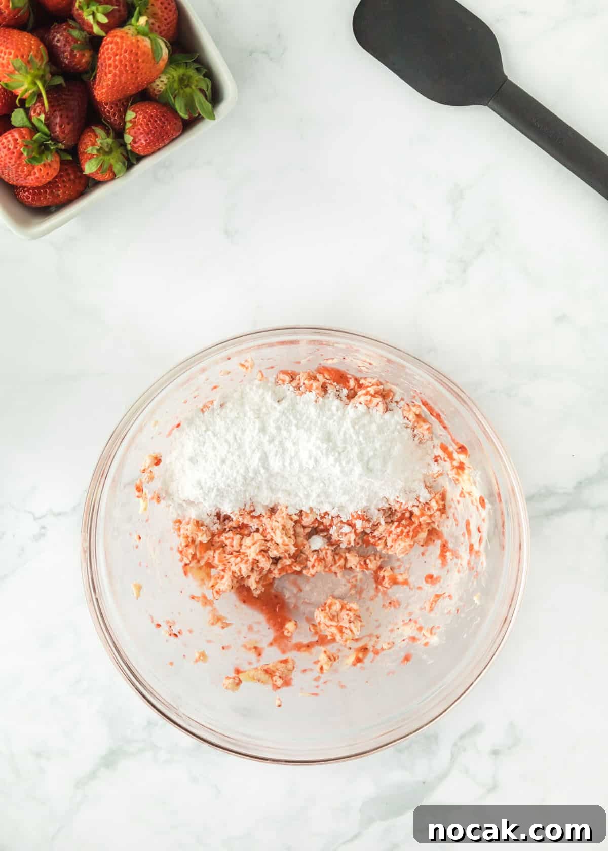 Strawberry Buttercream Frosting Recipe — Smooth, Fresh Flavor 7 Overhead photo of pureed strawberries with one full stick of butter and vanilla mixed together and powdered sugar not mixed together. The upper left and right corners are accented with whole strawberries and a black spatula.