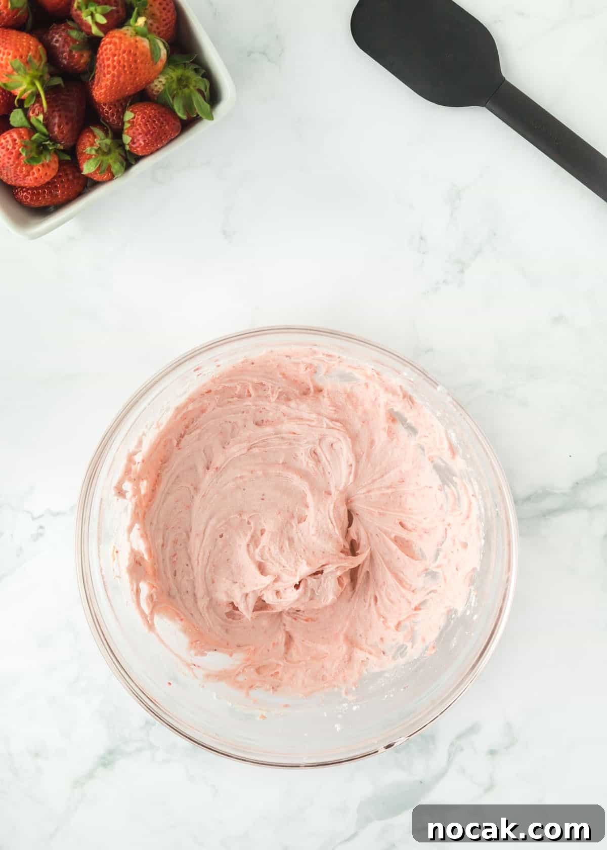 Strawberry Buttercream Frosting Recipe — Smooth, Fresh Flavor 8 Overhead photo of pureed strawberries with one full stick of butter, vanilla, and powdered sugar mixed together. The upper left and right corners are accented with whole strawberries and a black spatula.
