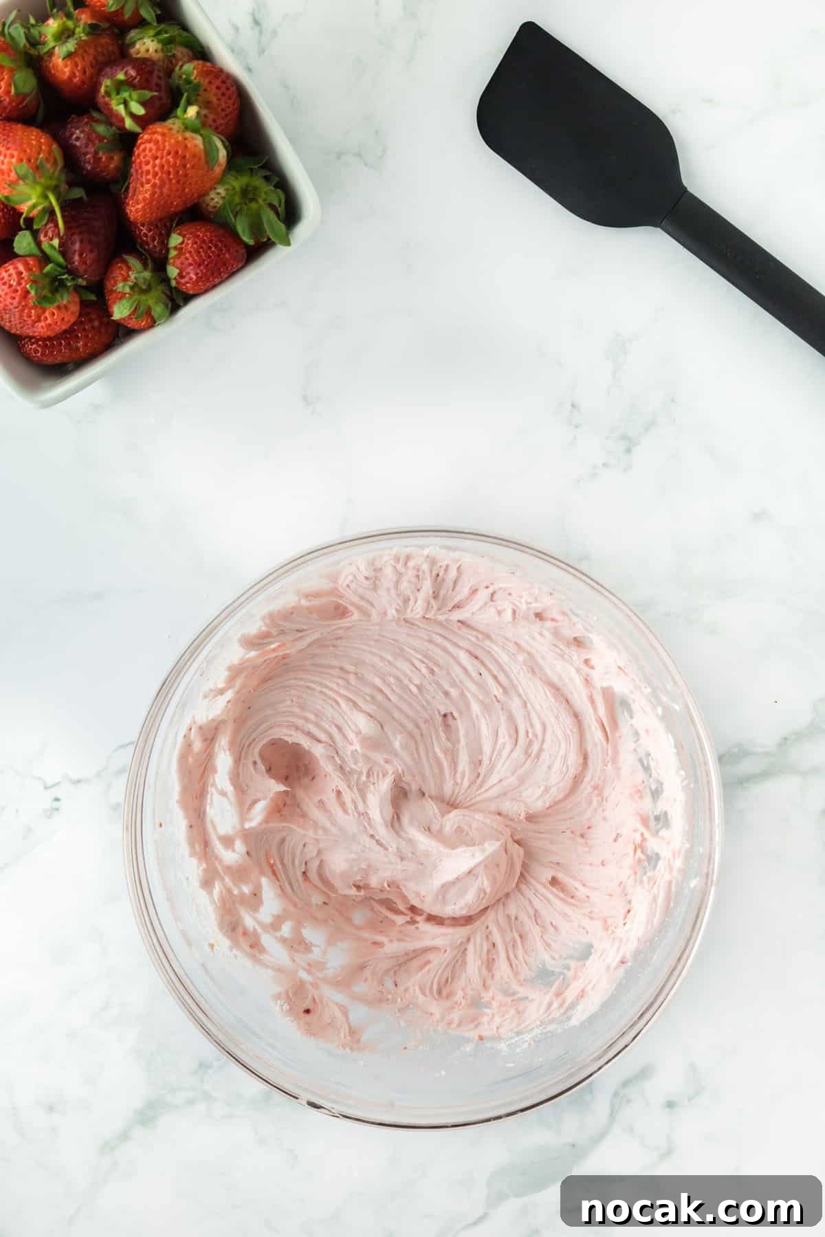 Strawberry Buttercream Frosting Recipe — Smooth, Fresh Flavor 10 Overhead photo of pureed strawberries with one full stick of butter, vanilla, powdered sugar, and heavy cream mixed together. The upper left and right corners are accented with whole strawberries and a black spatula.