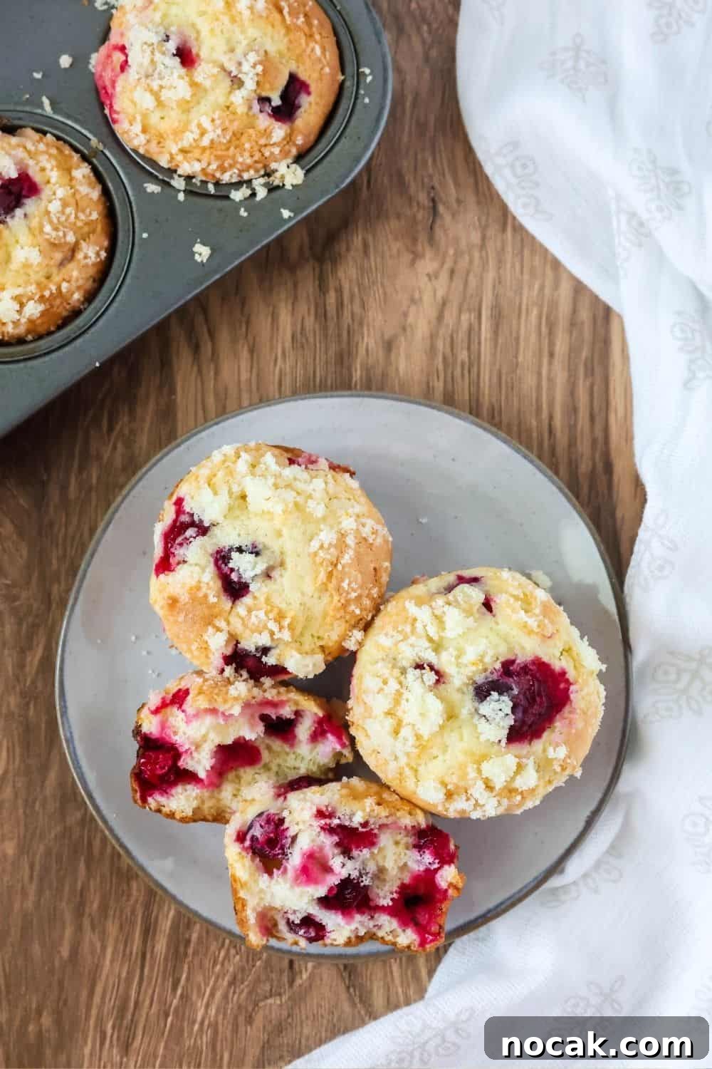 Cranberry Orange Muffins Made with Cake Mix 3 overhead view of easy cranberry orange muffins on an ivory-colored plate. One muffin has been torn in half to show all of the cranberries inside. A muffin pan is in the background.