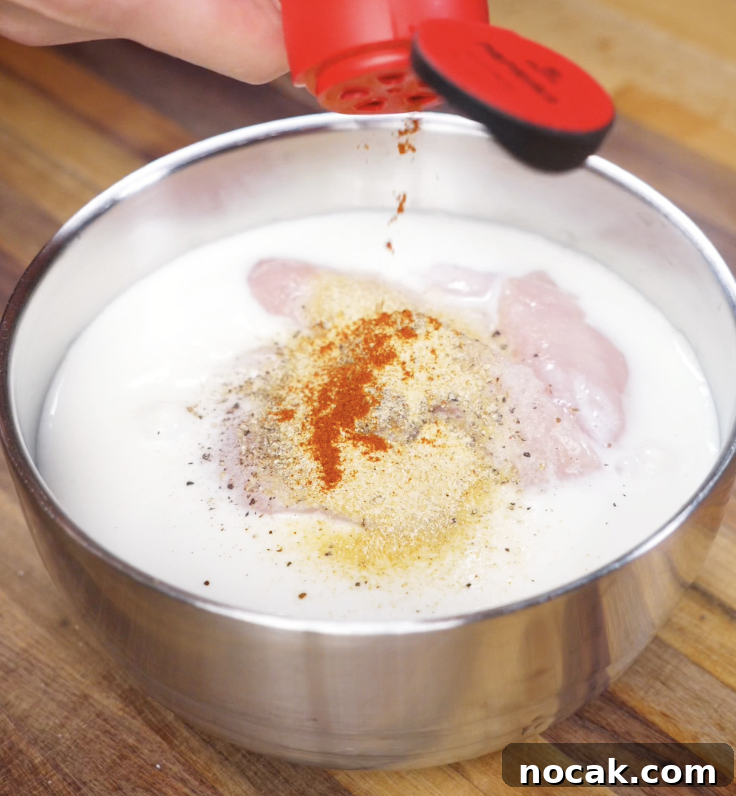 Crispy Garlic Parmesan Chicken Tenders Recipe 4 Paprika and garlic powder are sprinkled into a bowl of buttermilk and raw chicken tenders, creating the flavorful marinade base.