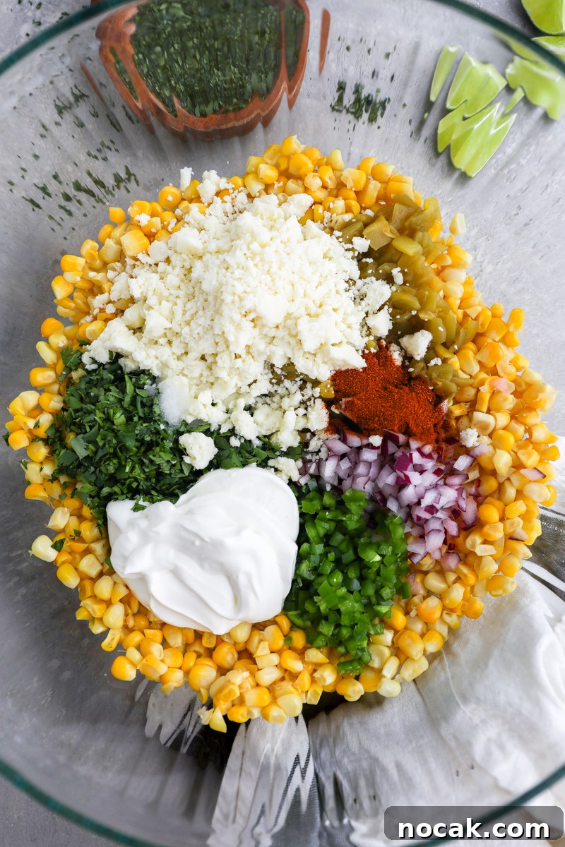 Mexican Street Corn Dip Recipe: Creamy Elote-Inspired Dip 4 A large clear mixing bowl filled with cooked corn, green chiles, red onion, jalapeno, sour cream, cilantro, cotija cheese, chili powder, salt and lime juice