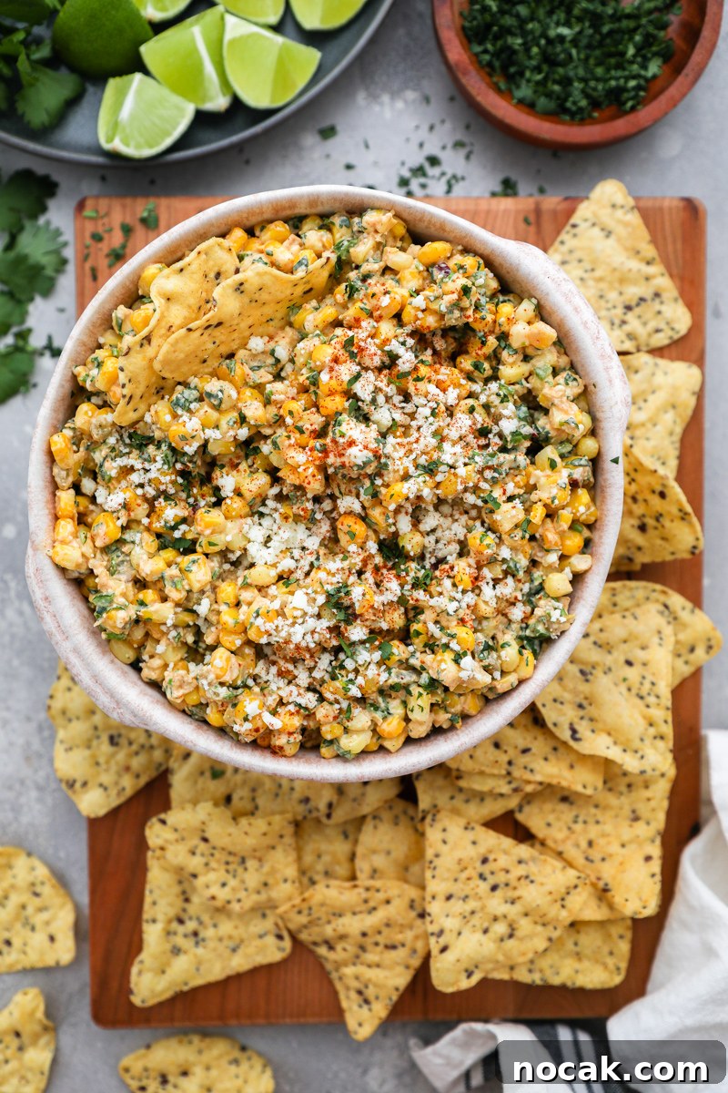 Mexican Street Corn Dip Recipe: Creamy Elote-Inspired Dip 7 A bowl of healthier Mexican street corn dip sitting on a wood board next to tortilla chips, lime wedges and a small bowl of chopped cilantro