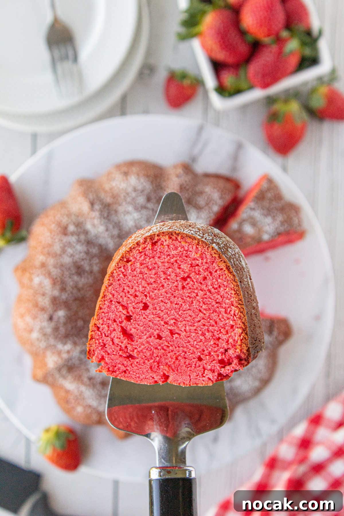 Old-Fashioned Strawberry Pound Cake with Cream Cheese Frosting 2 A cake server holds a slice of strawberry bundt cake over the sliced bundt cake and strawberries on marble counter and plate below.