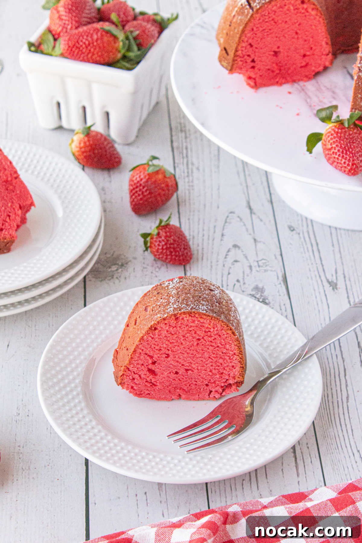 Old-Fashioned Strawberry Pound Cake with Cream Cheese Frosting 6 A piece of sliced red strawberry bundt cake on a white plate with a fork ready to serve as strawberries and cut cake are displayed behind it.
