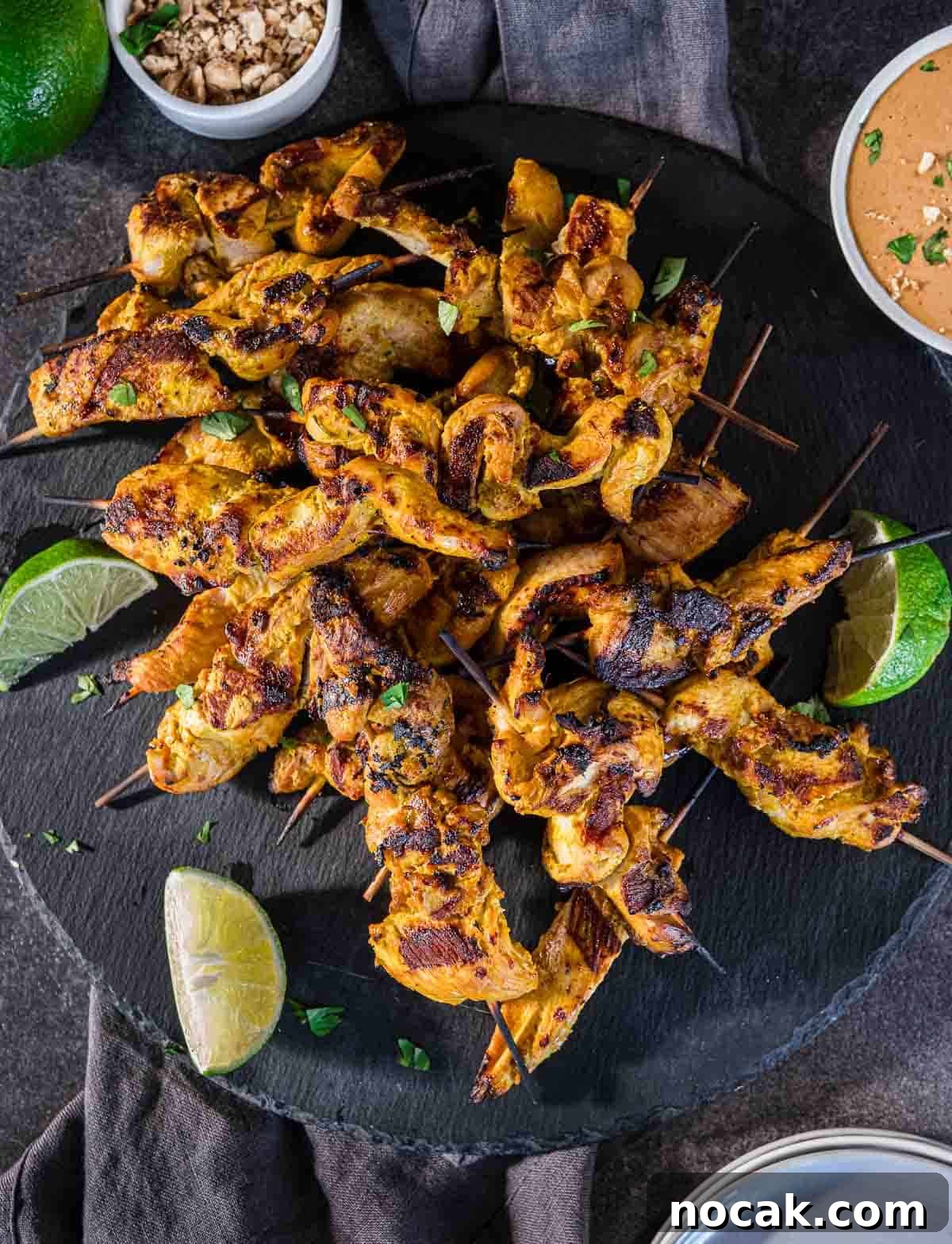 Chicken Satay Skewers with Creamy Peanut Dipping Sauce 2 Grilled chicken satay skewers on a slate board surrounding by lime wedges.