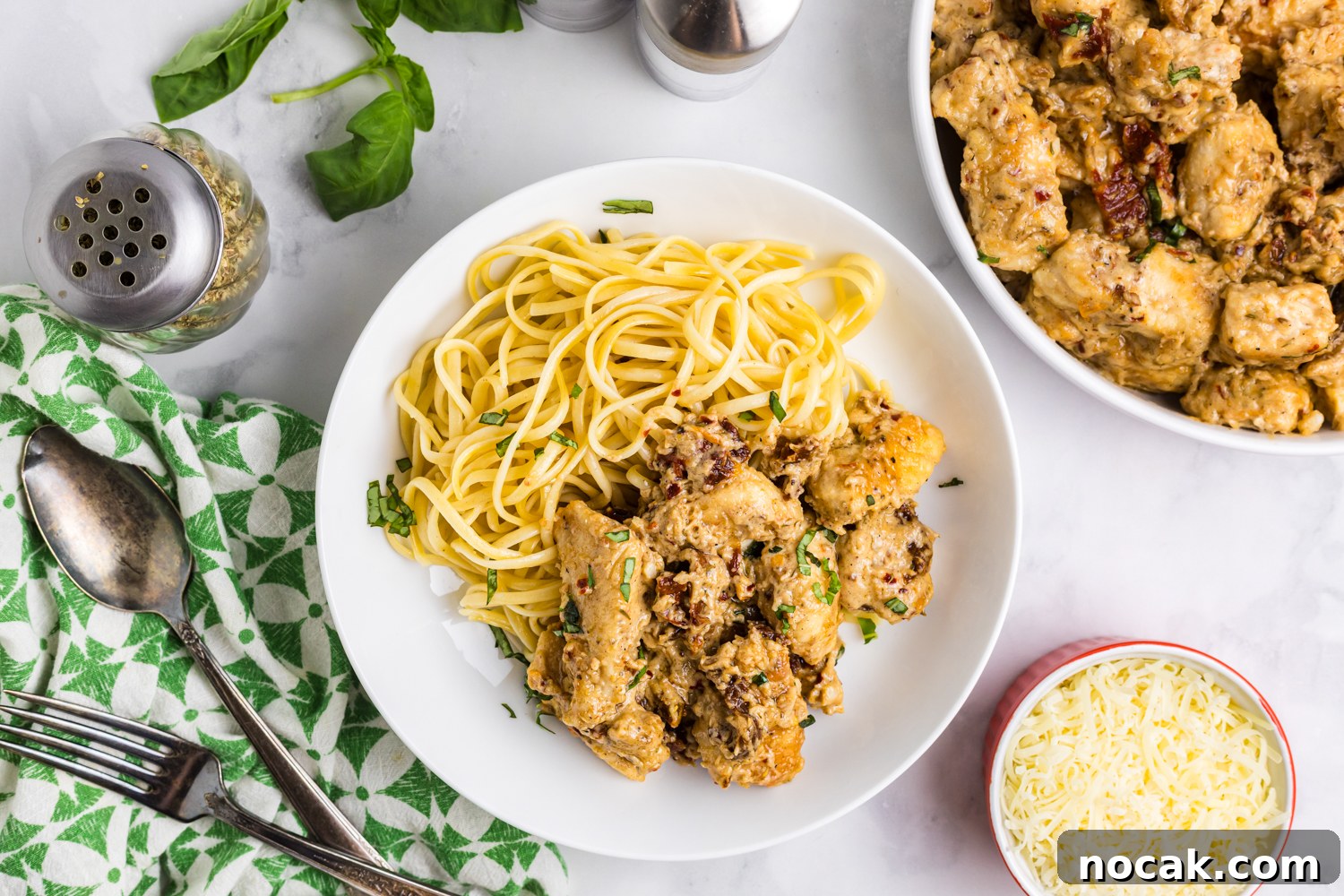 Blackstone Marry Me Chicken Recipe: Skillet Chicken with Creamy Sun-Dried Tomato Sauce 8 Overhead view of a bowl of pasta topped with creamy sun-dried tomato chicken, garnished with herbs and surrounded by utensils, cheese, basil leaves, and seasonings on a light countertop.