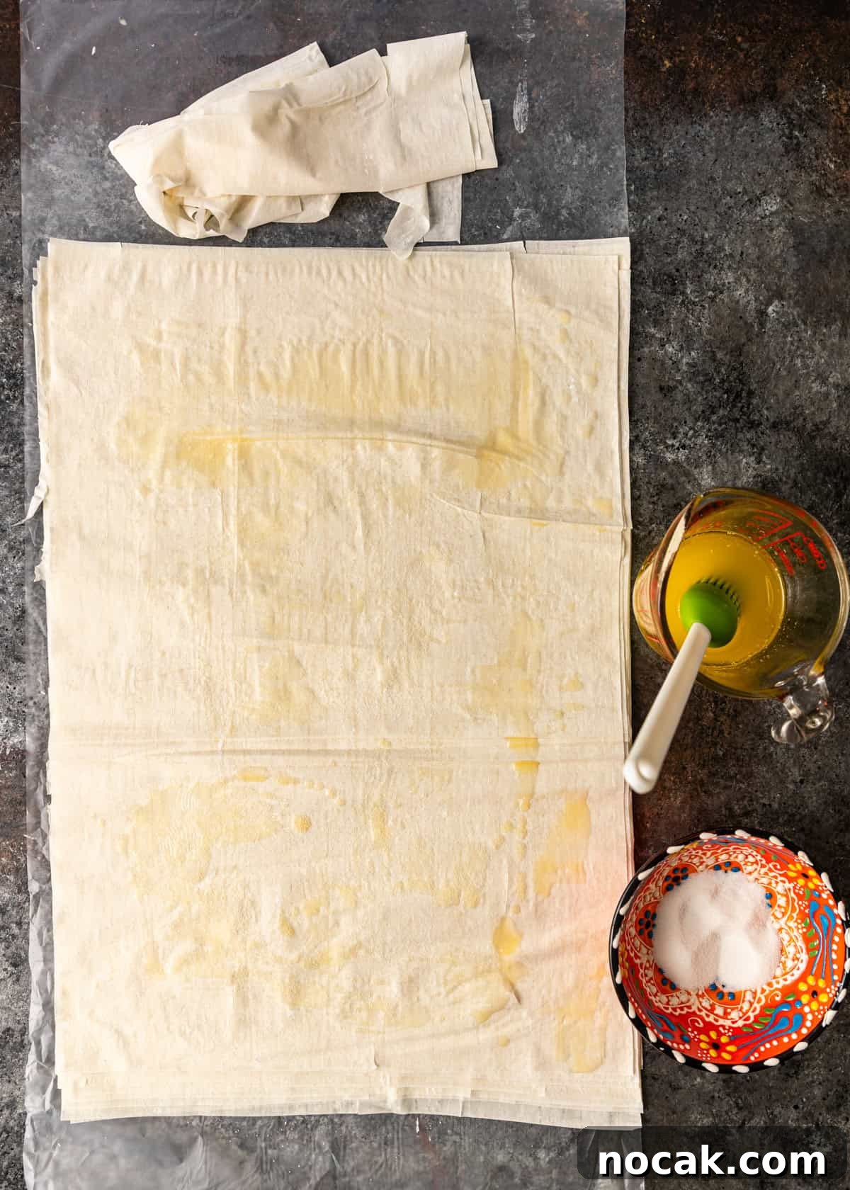 Baklava Cheesecake Bars with Honeyed Pistachios 5 overhead process shot: phyllo dough on wax paper with measuring cup of butter off to side and bowl of sugar