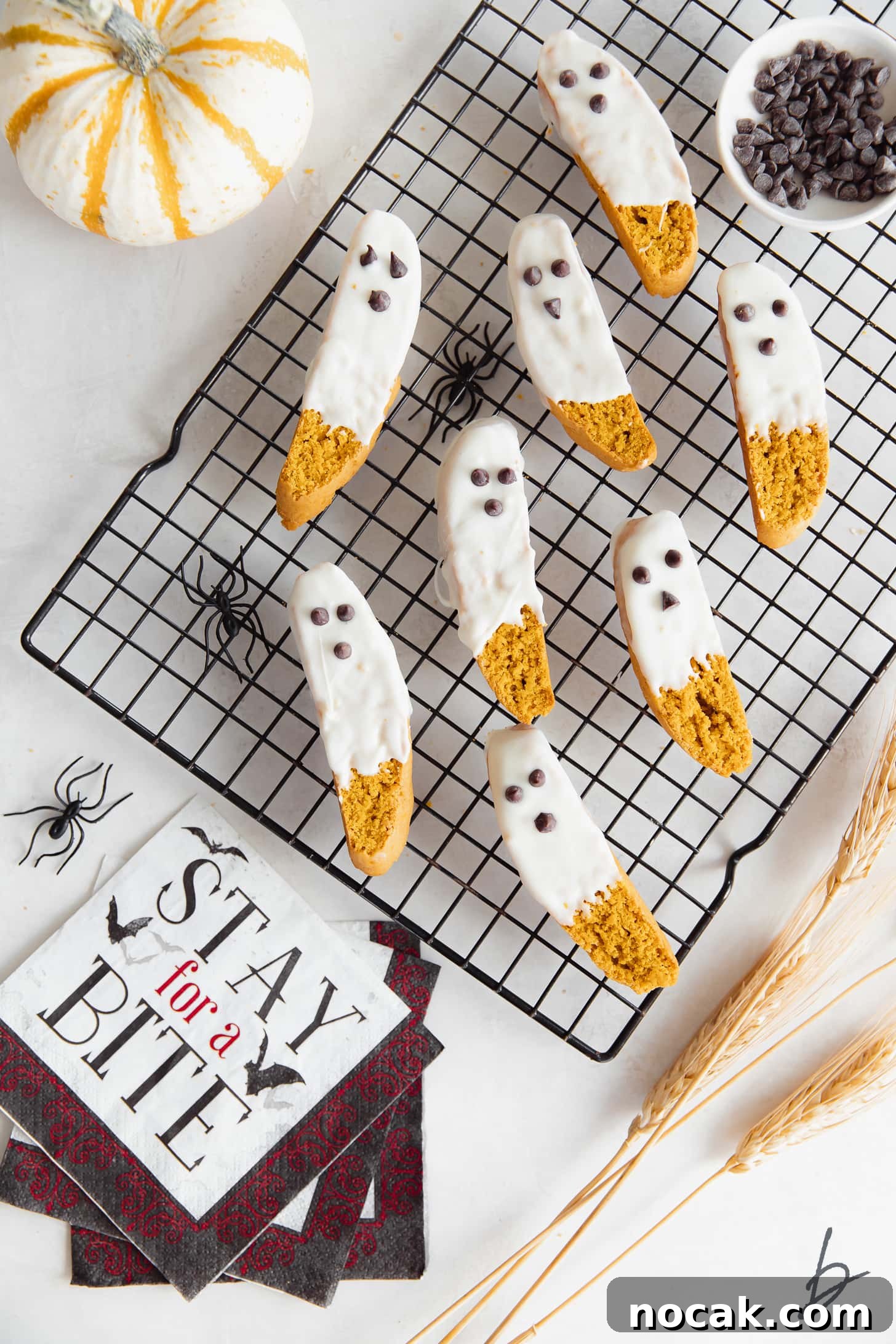 Pumpkin Spice Biscotti Recipe: Crunchy Autumn Cookie Guide 5 White chocolate dipped pumpkin biscotti that look like ghosts on black wire cooling rack next to halloween napkin and mini pumpkin.