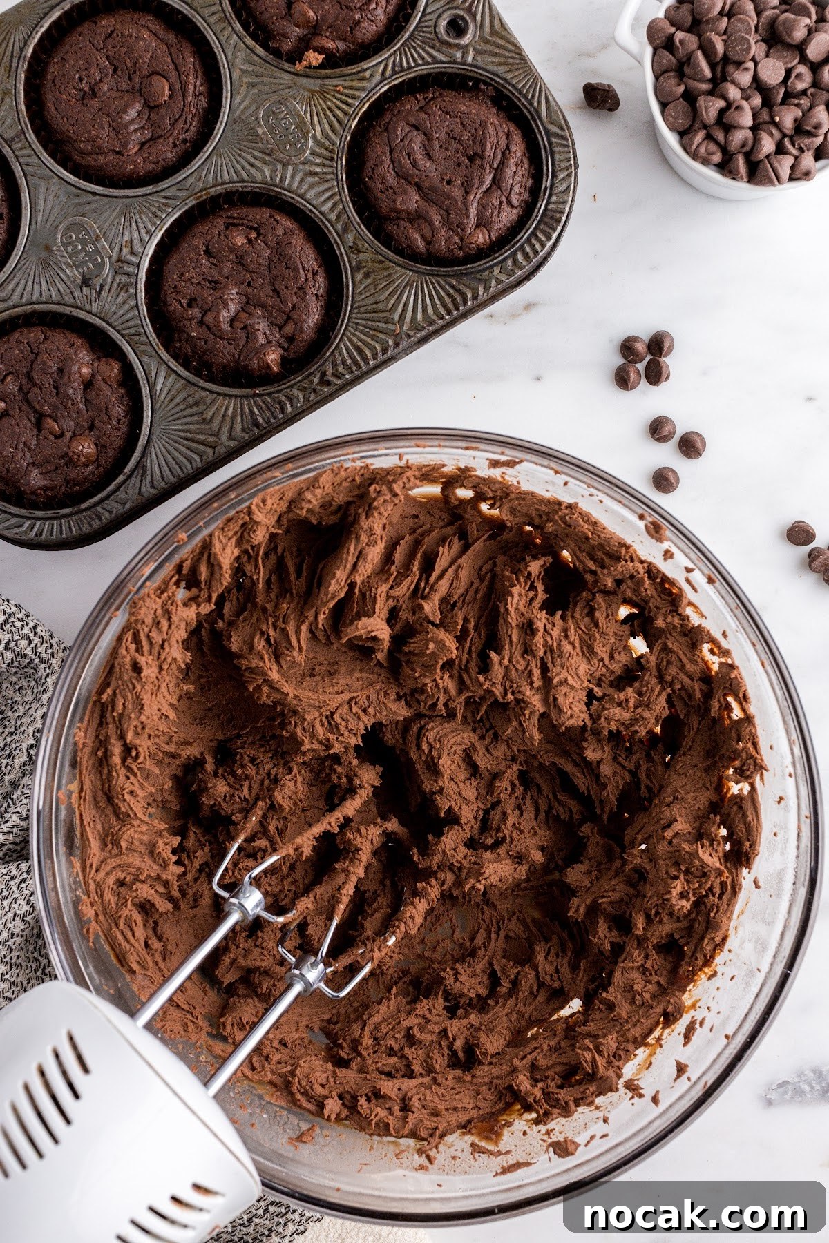 Fudgy Brownie Cupcakes with Gooey Chocolate Centers 12 Mixing the frosting in a bowl with a handheld mixer until stiff peaks form, with cupcakes in the background.
